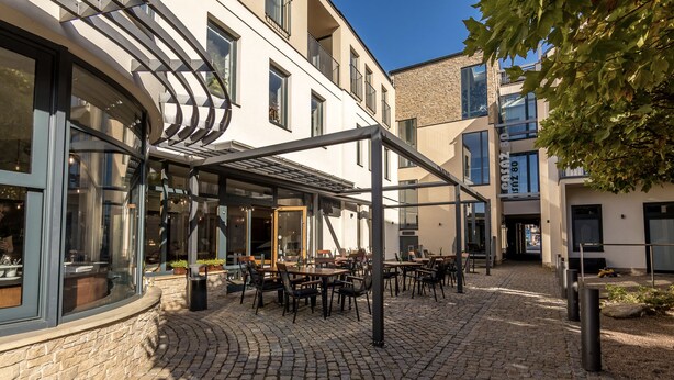 Un espace café en plein air avec des tables et des chaises, un chemin pavé et un bâtiment doté de grandes fenêtres et d’une façade en pierre.
