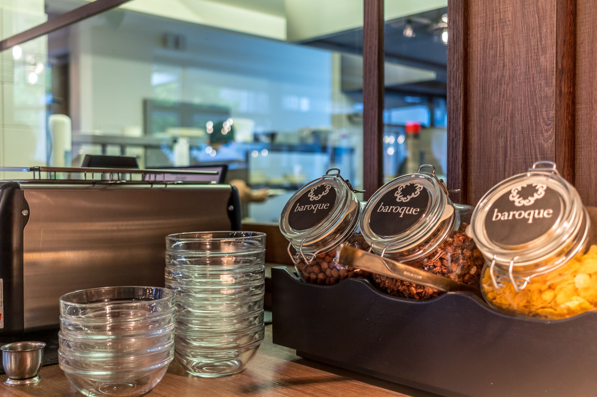 Uma área de bar com uma máquina de café, copos empilhados e potes com o rótulo 'baroque' contendo lanches.