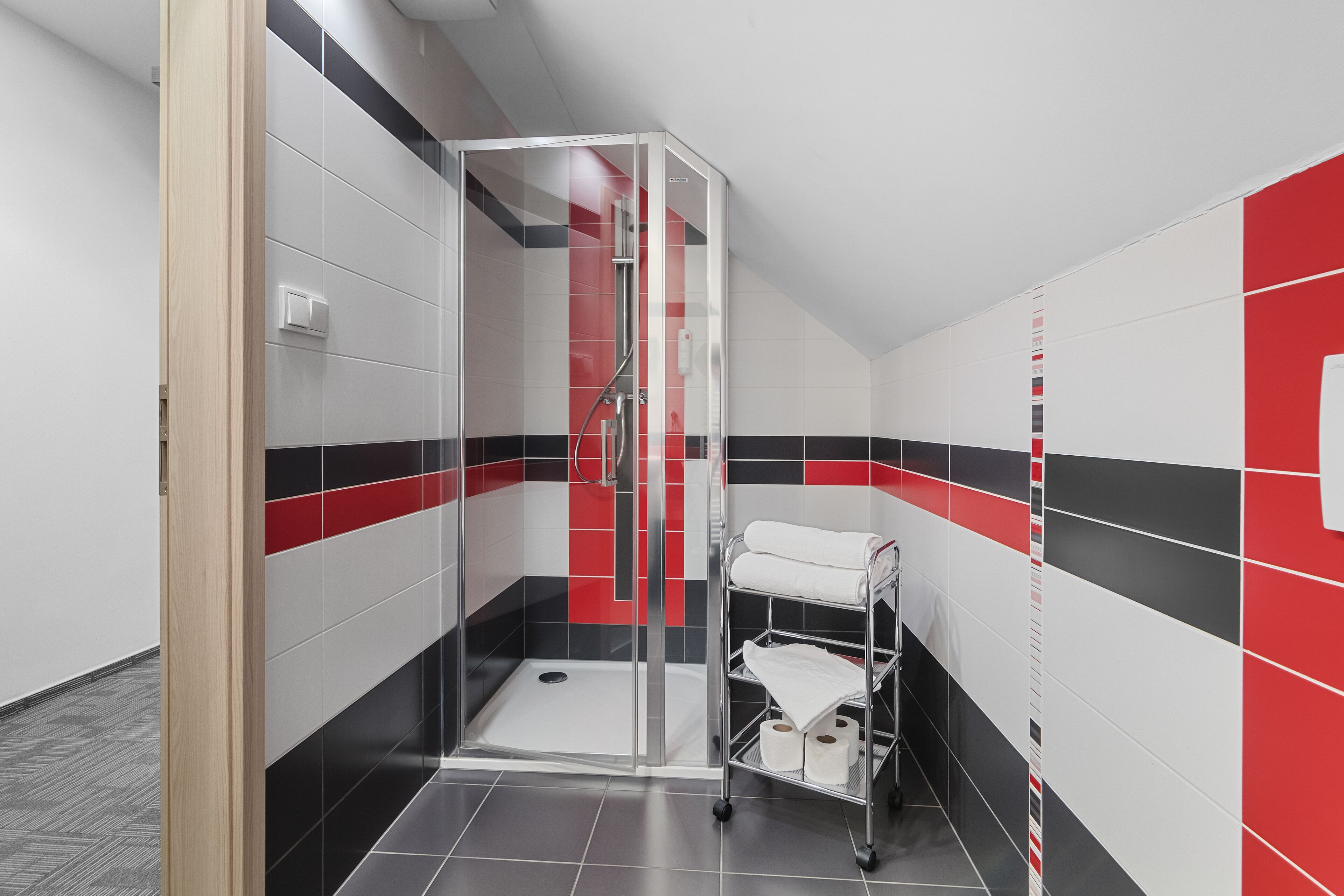 Un baño moderno con cabina de ducha de vidrio, toallero blanco y pared con mosaicos rojos y negros.