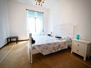 Quarto com cama, mesa de cabeceira, uma pequena mesa e janela com cortinas.