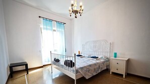 Een slaapkamer met een bed met wit metalen frame, een nachtkastje, een bankje en een raam met gordijnen.