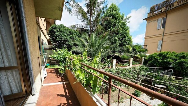 Een balkon met een metalen hekwerk, betegelde vloer en uitzicht op een tuin met verschillende planten en bomen.