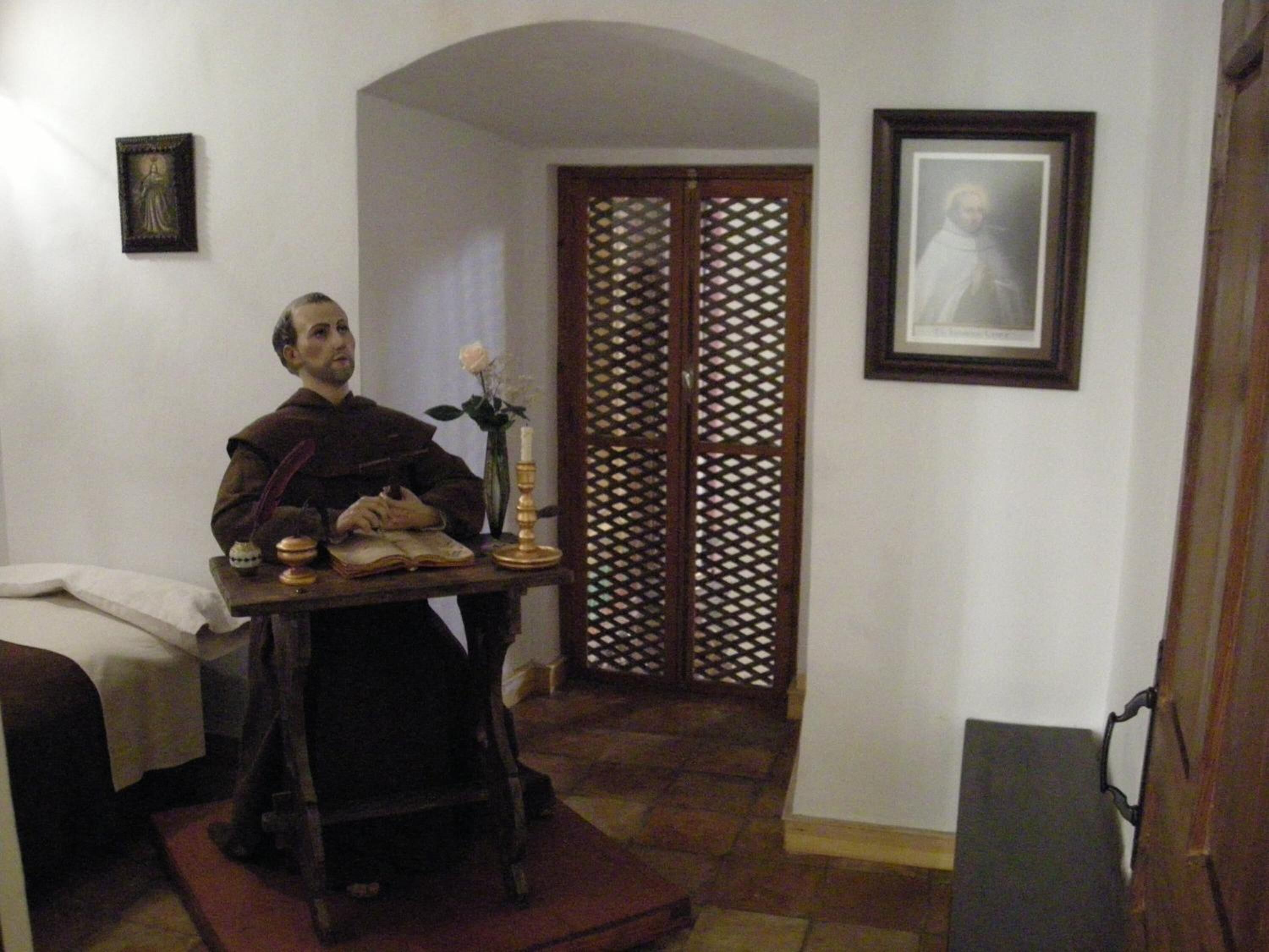 A room with a bed, a table with a book and a candle, a wooden door, and a framed picture on the wall.