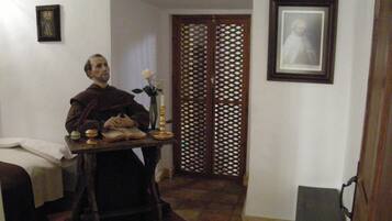 A room with a bed, a table with a book and a candle, a wooden door, and a framed picture on the wall.