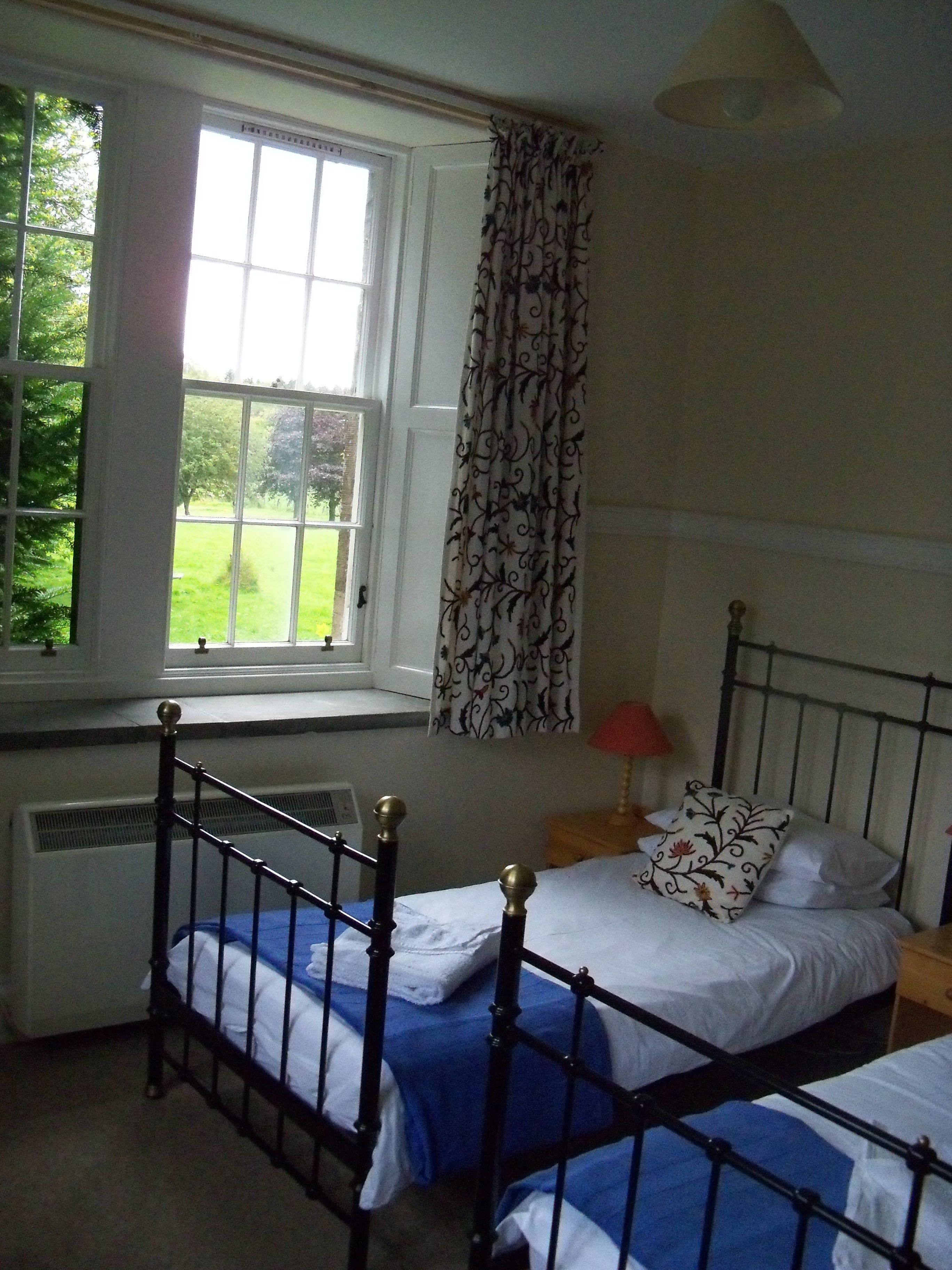 A bedroom with two beds, a window with curtains, a radiator, and a nightstand with a lamp.