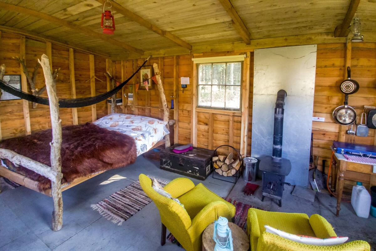 A rustic cabin interior with a bed, yellow chairs, a fireplace, and a dining area.