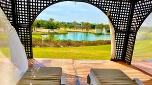 A view from a covered outdoor seating area with a pool and greenery in the background.