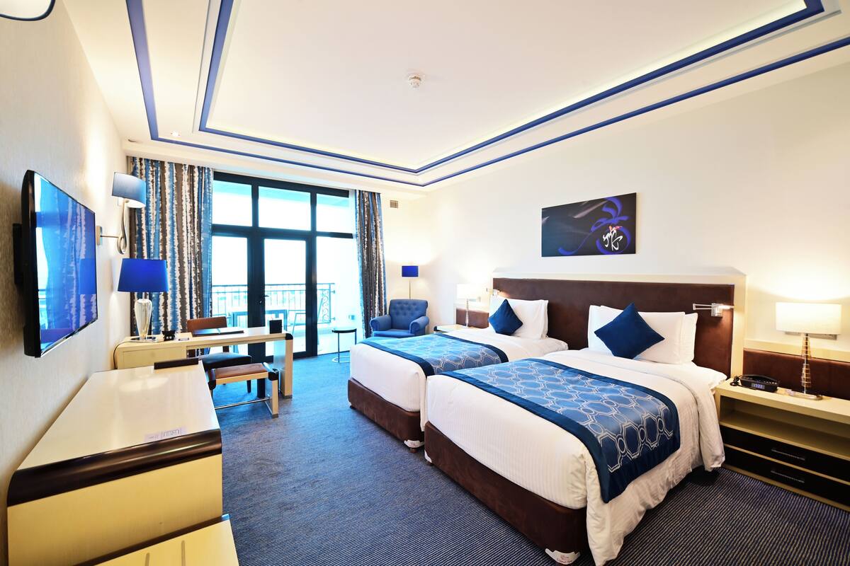 Ein Hotelzimmer mit zwei Betten, einem Schreibtisch, einem Stuhl, einem Fernseher und einem Balkon mit Vorhängen.