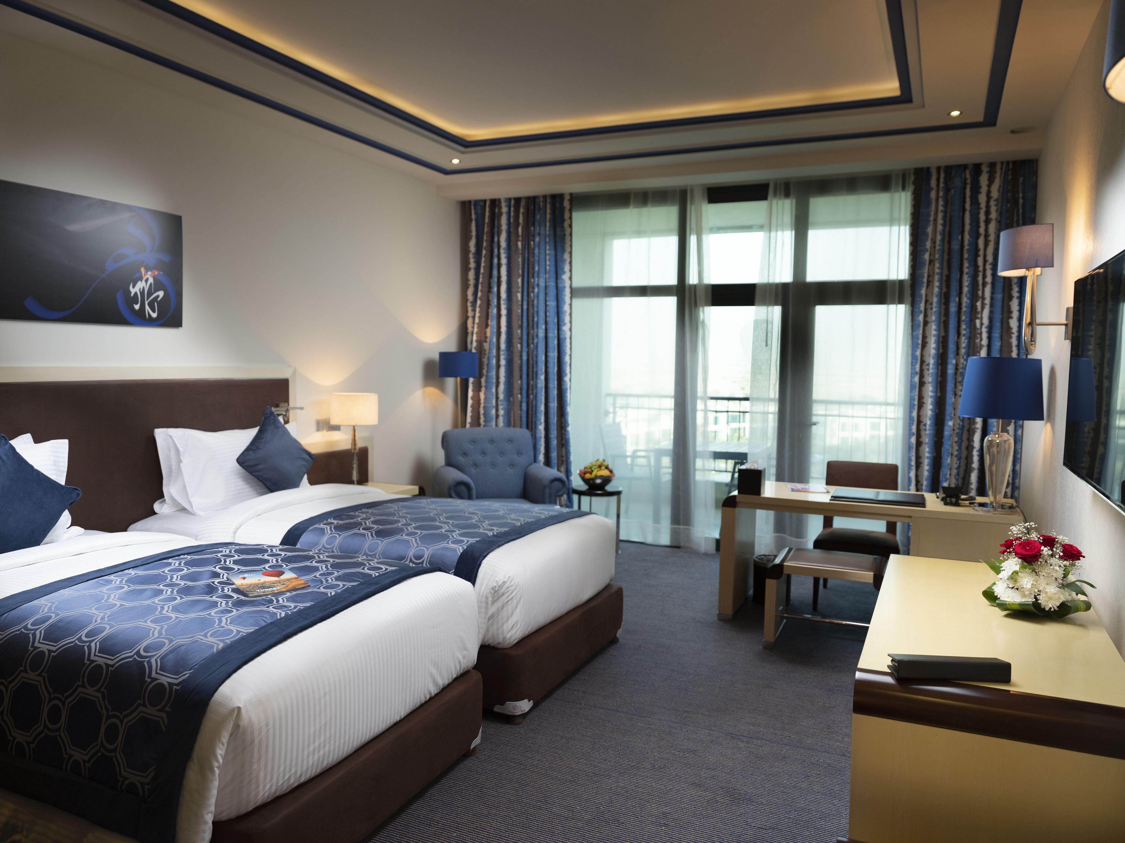 Ein Hotelzimmer mit zwei Betten, einem Schreibtisch, einem Sessel, einem Fernseher und einem Balkon mit Vorhängen.