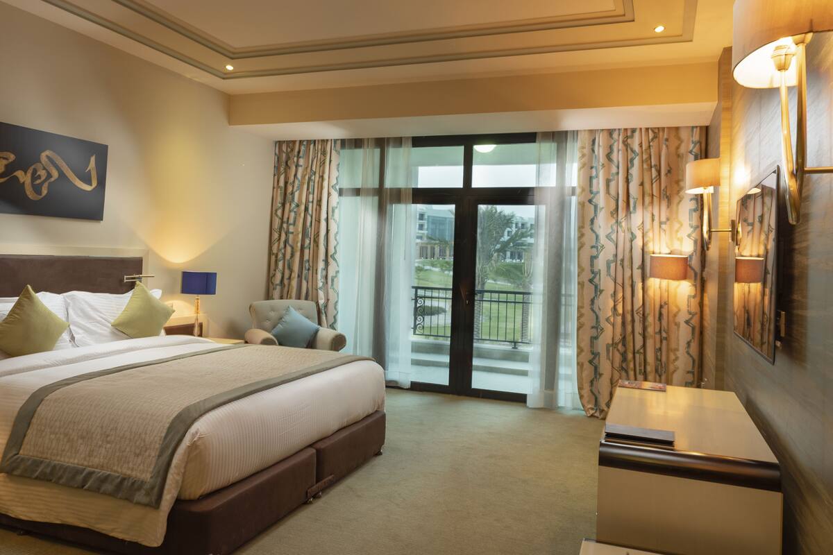 Ein Hotelzimmer mit einem großen Bett, einem Nachttisch, einer Lampe und Blick ins Freie durch das Fenster.