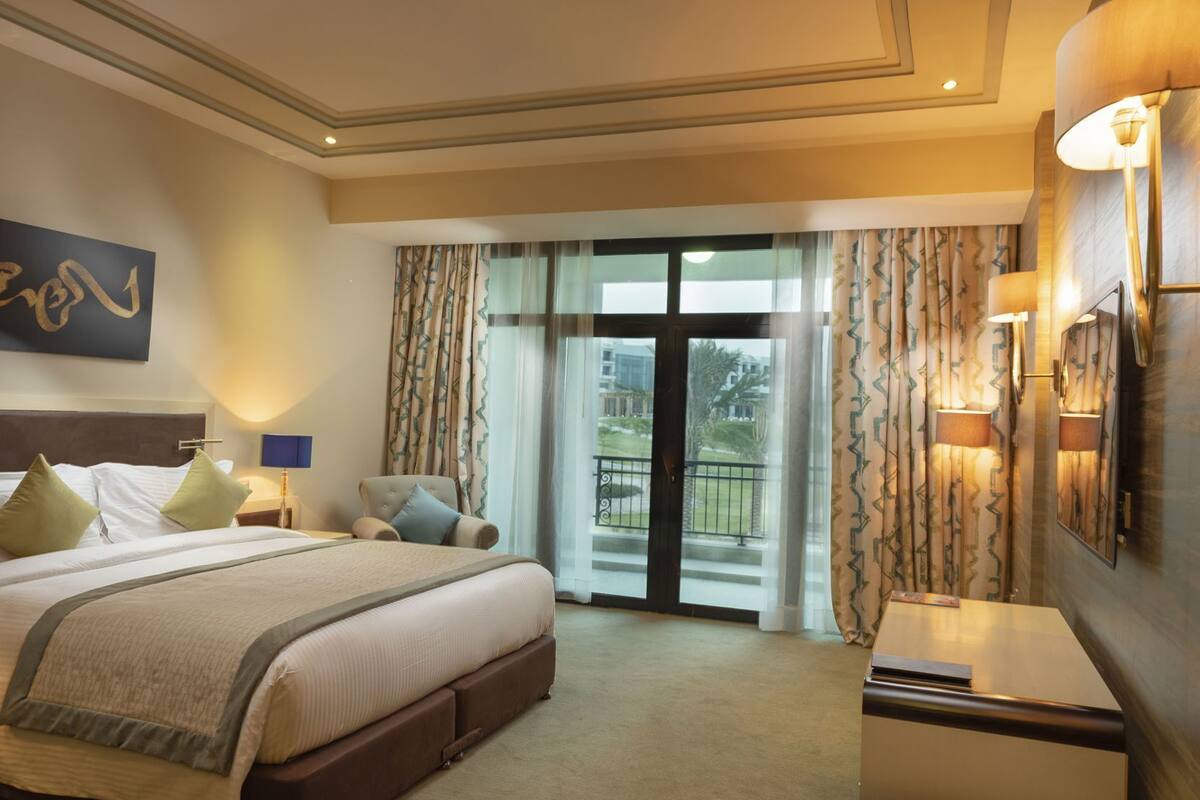Ein Hotelzimmer mit einem großen Bett, einem Nachttisch, einer Lampe, Blick ins Freie durch eine Glastür und einer Wandleuchte.