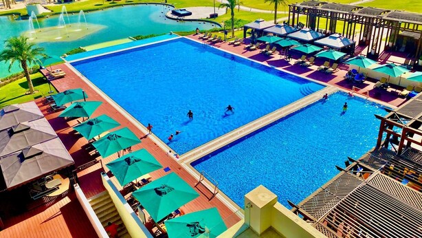 A large swimming pool with multiple lanes, surrounded by sun loungers and umbrellas, with a view of a resort area in the background.