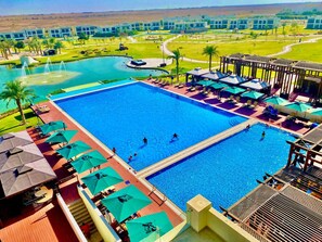 Une grande piscine avec plusieurs couloirs de nage, entourée de chaises longues et de parasols, et offrant une vue sur un complexe hôtelier en arrière-plan.