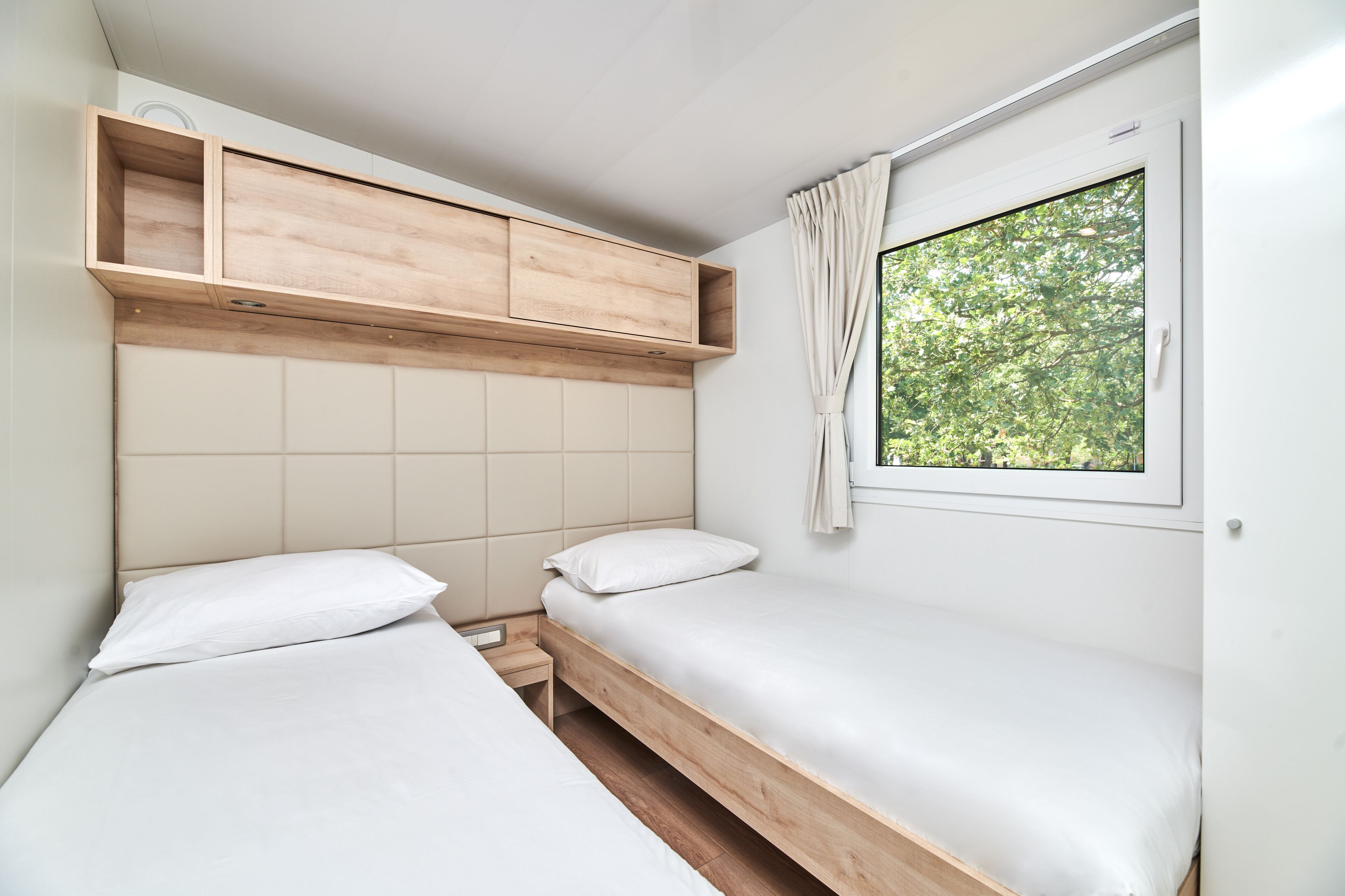 Una habitación pequeña y minimalista con dos camas individuales, cabecera de madera, estante empotrado y ventana con vista a árboles.