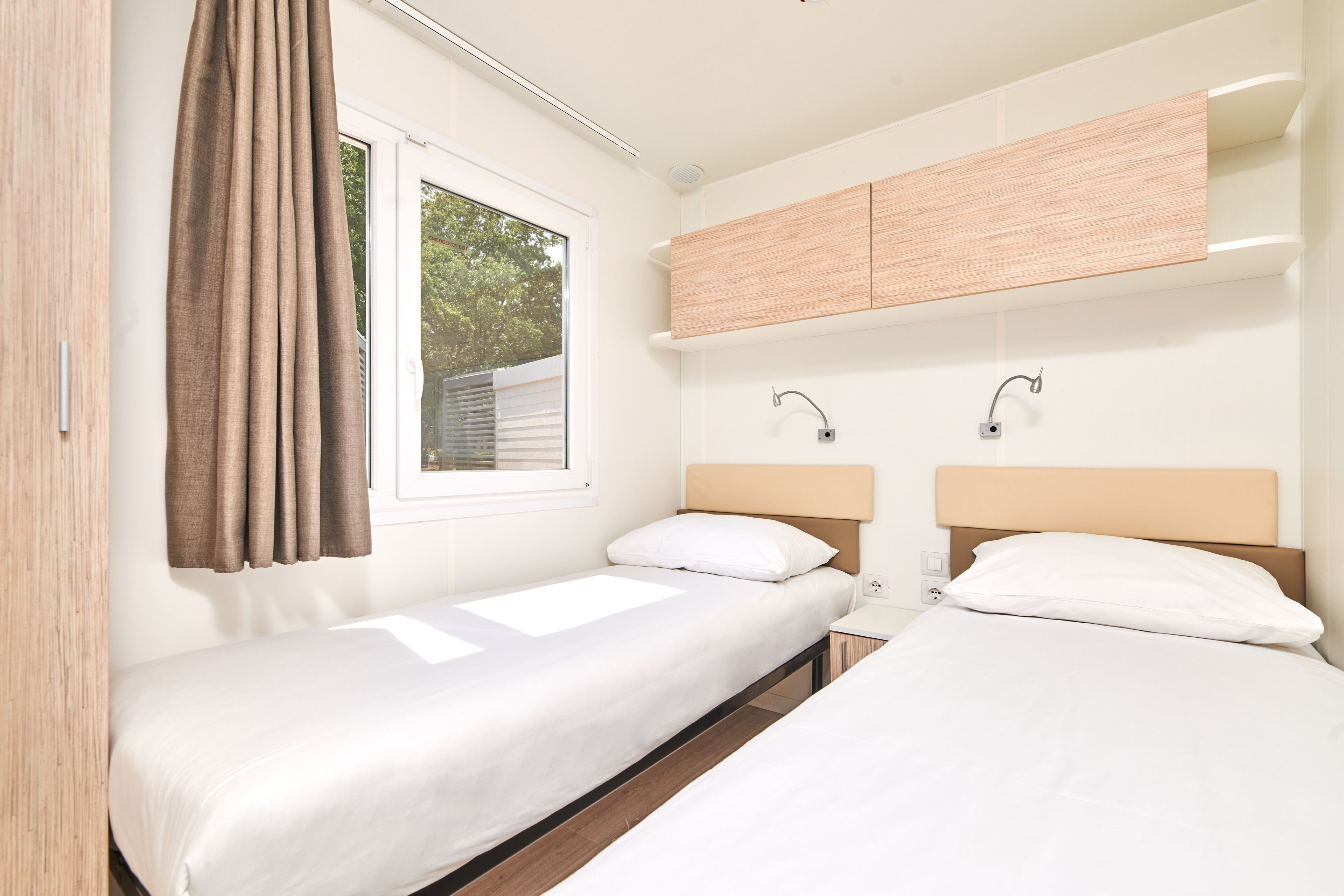 Een slaapkamer met twee enkele bedden, een raam met gordijnen, een houten hoofdeinde en een ingebouwd plankje boven de bedden.