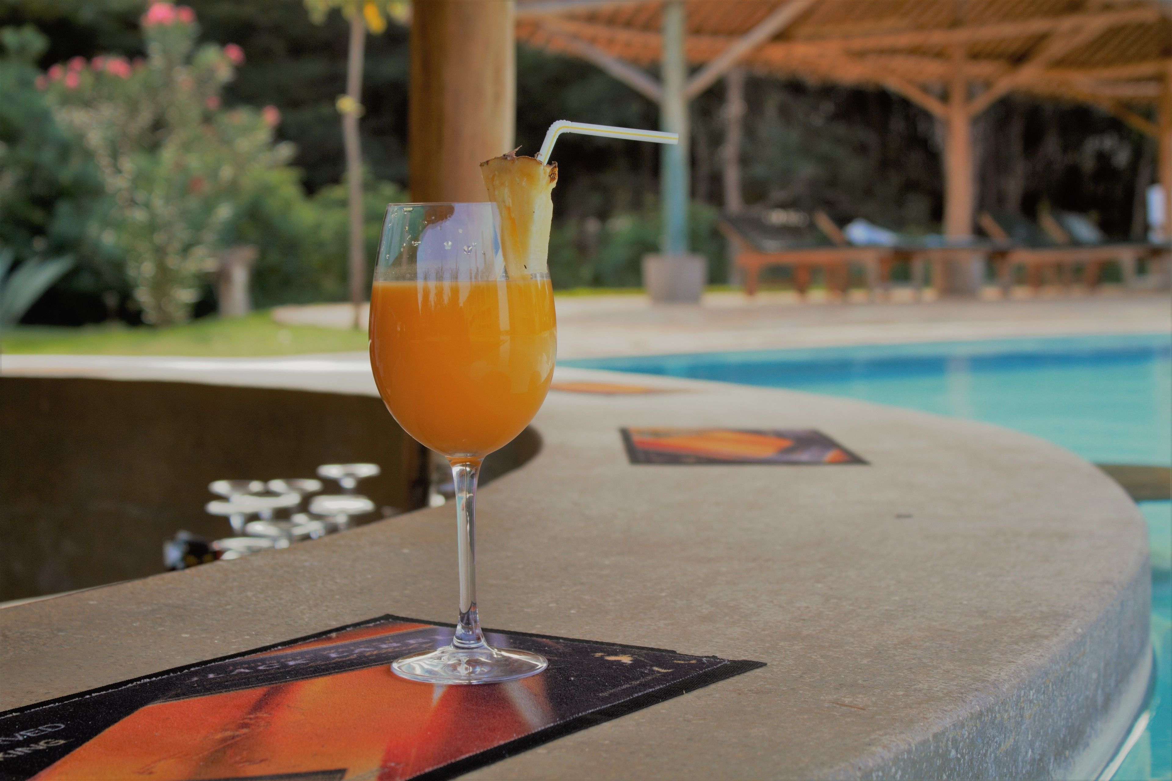 A glass of orange juice with a straw and a slice of pineapple on a table by a pool.