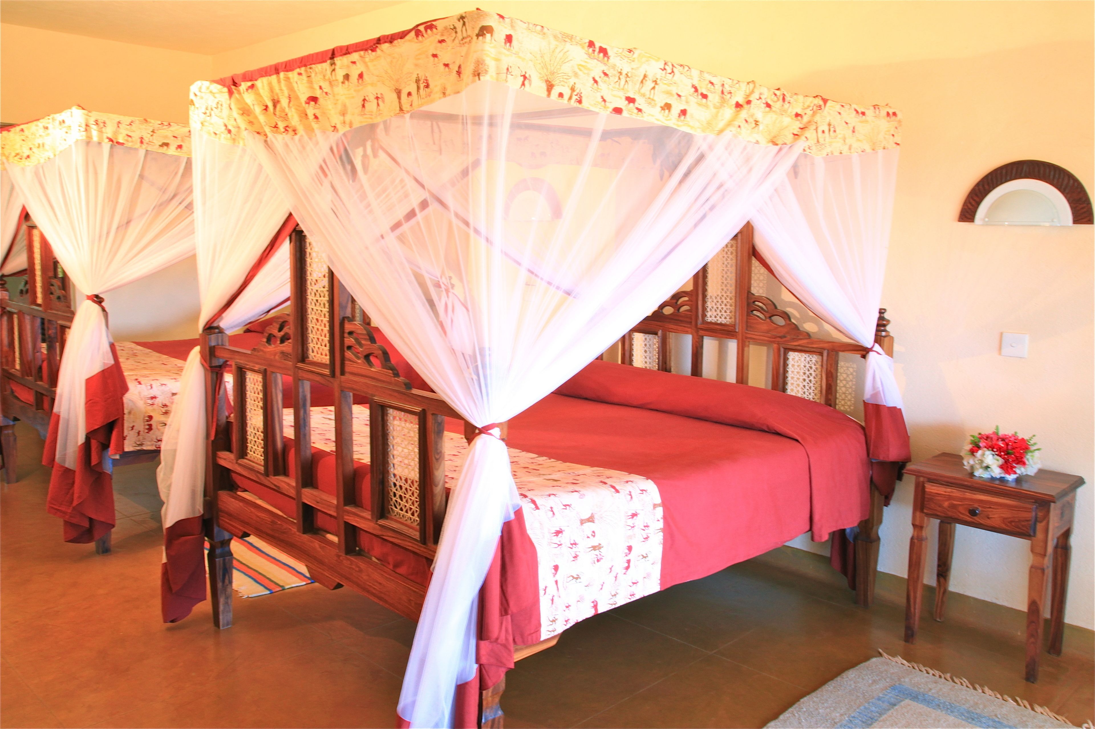A room with four wooden beds, each with a canopy bedspread and a red bed skirt. There is a wooden nightstand with a vase of flowers beside one bed.