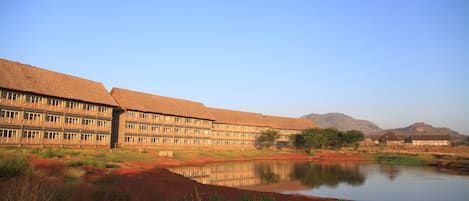 A large building with multiple balconies, a reflective water body in the foreground, and a mountainous backdrop.
