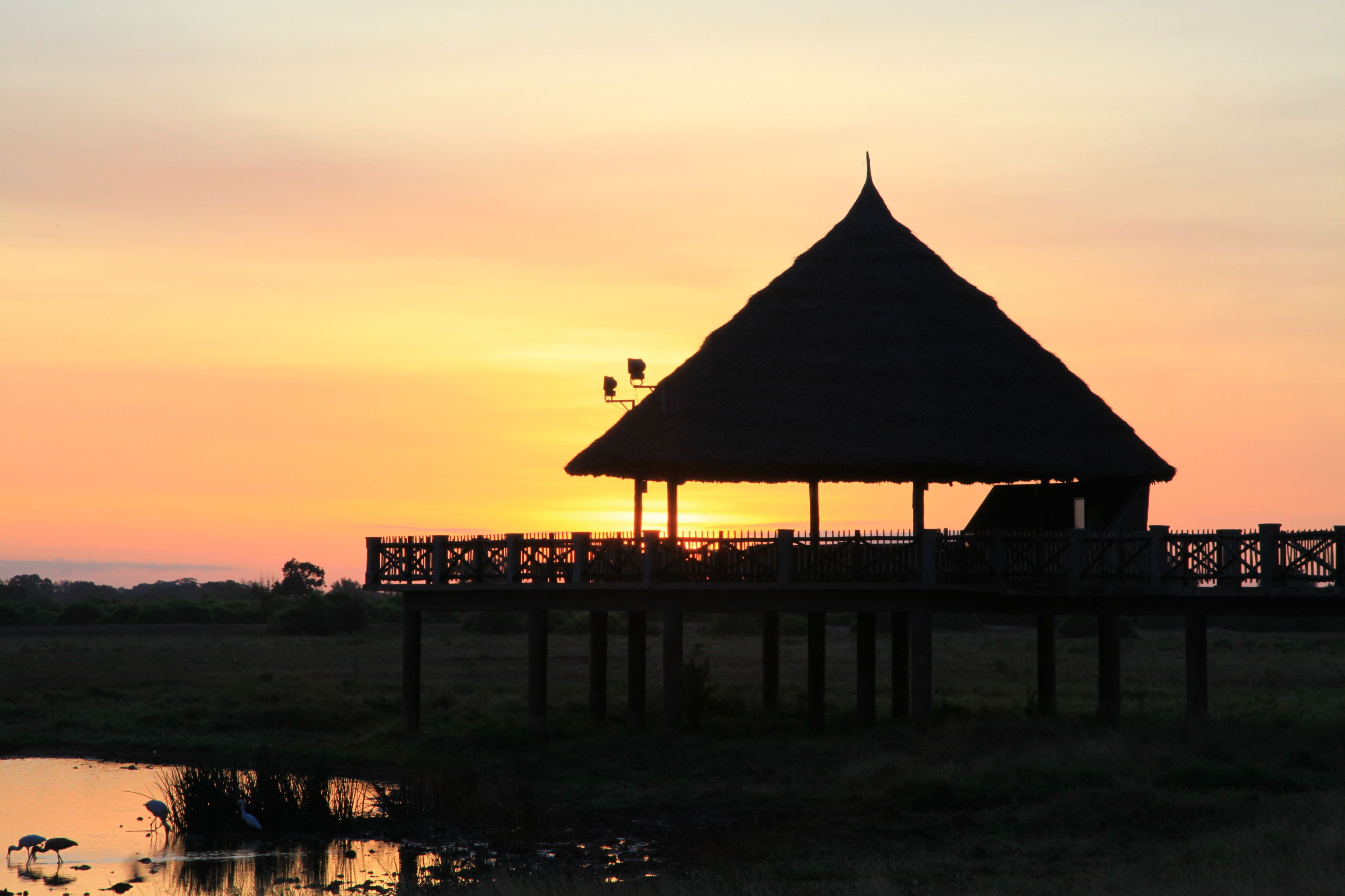 A thatched-roof pavilion on a pier over water, with a sunset in the background.