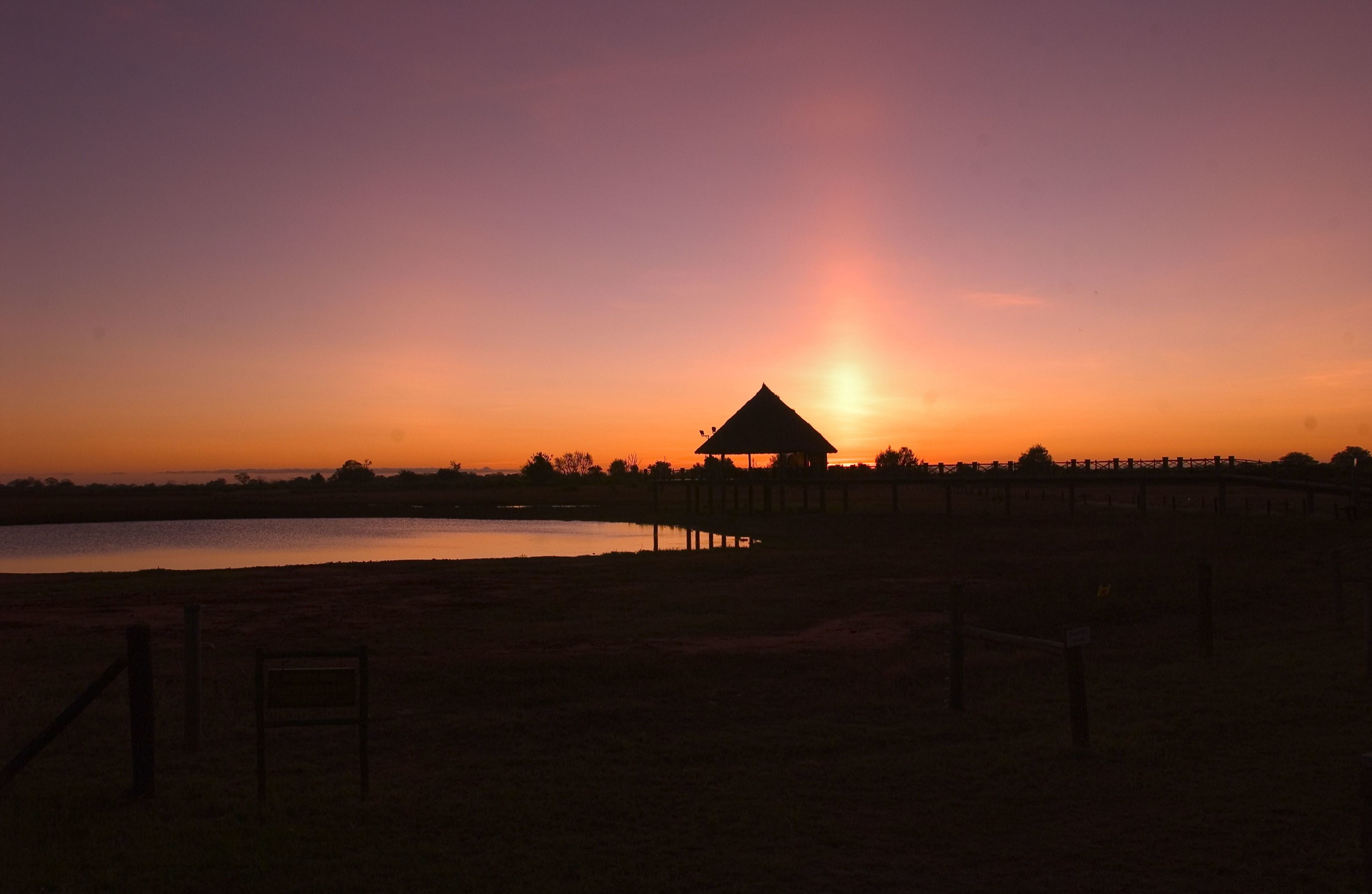 A sunset view with a thatched-roof structure near a body of water, surrounded by a fence and a few benches.