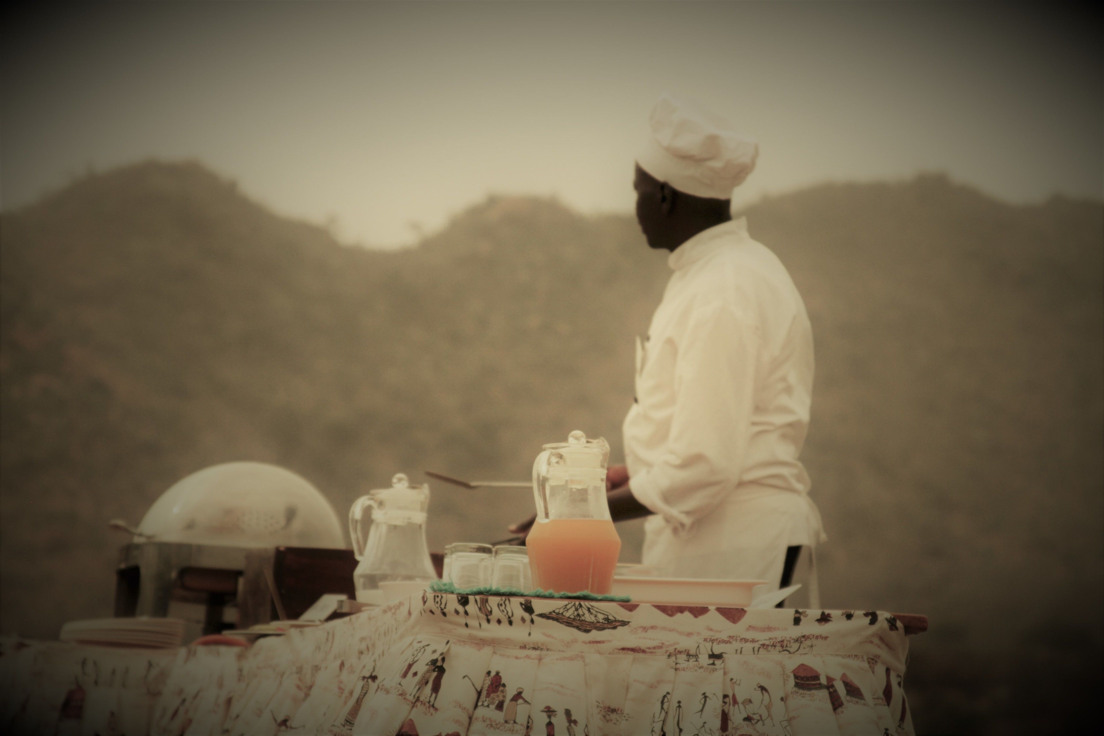 Un chef con uniforme blanco preparando comida en una mesa al aire libre con mantel estampado, una jarrón y una tetera.