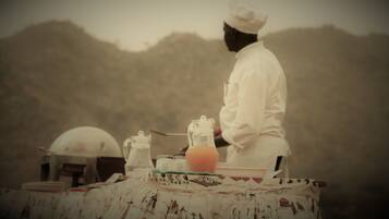 Un chef con uniforme blanco preparando comida en una mesa al aire libre con mantel estampado, una jarrón y una tetera.