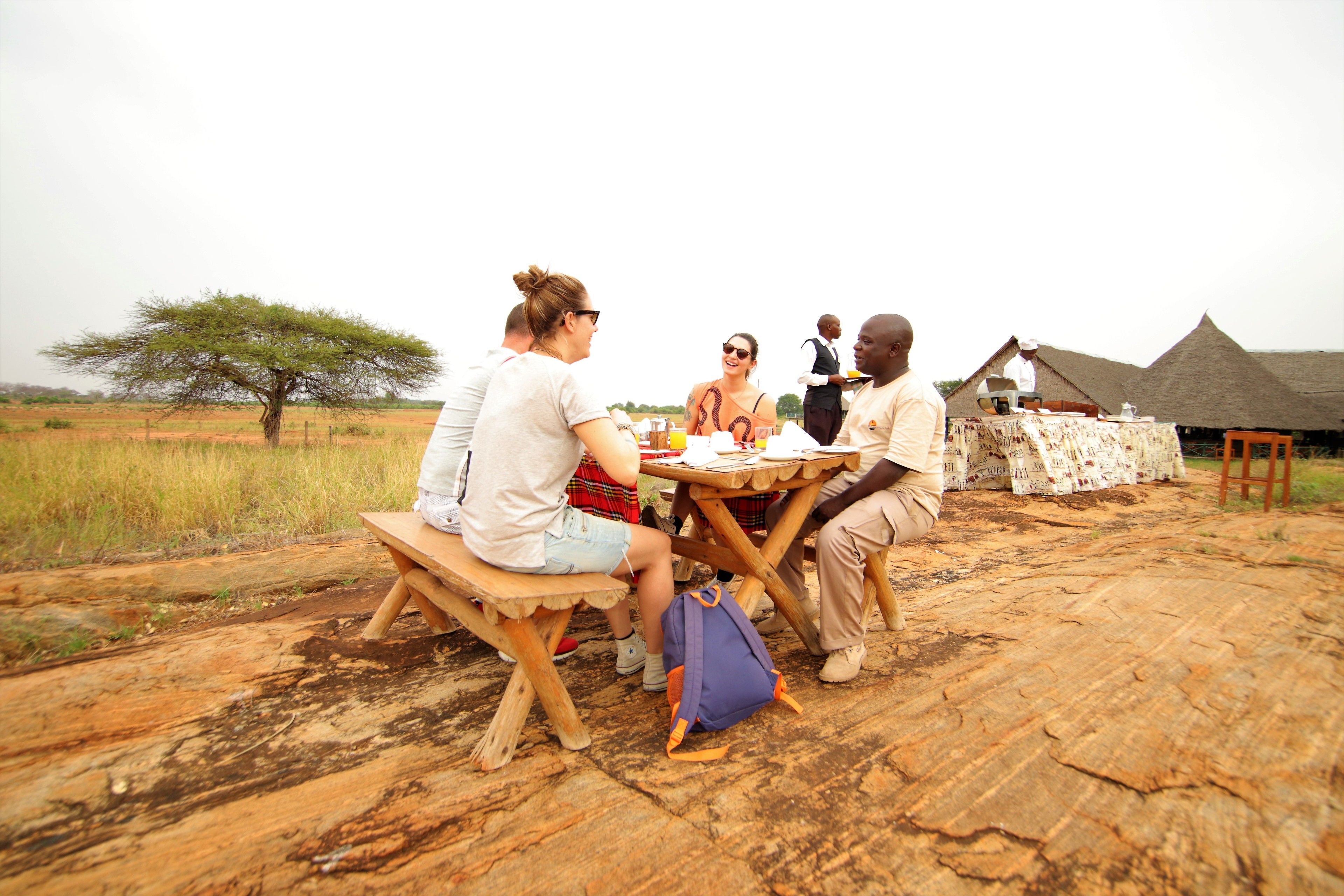 Un grupo de personas cenando al aire libre en una mesa de madera, con un edificio de techo de paja al fondo.