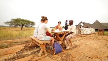 Un grupo de personas cenando al aire libre en una mesa de madera, con un edificio de techo de paja al fondo.