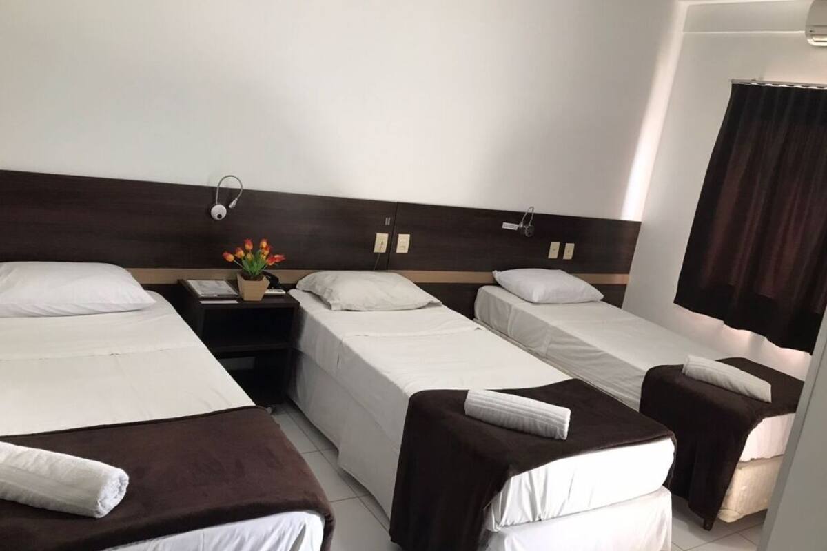 Habitación de hotel con tres camas individuales, una mesita de noche con lámpara y una pequeña mesa con un florero.
