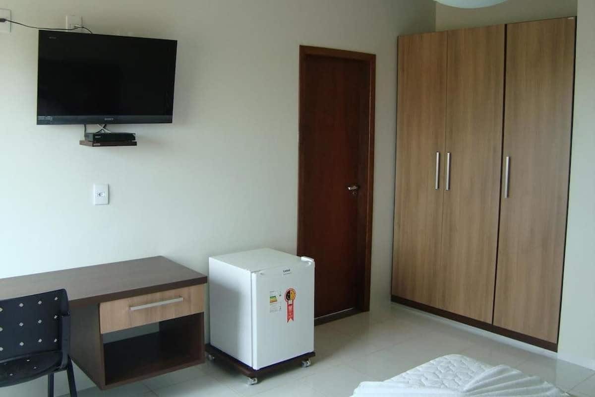 Una habitación con televisión de pantalla plana, un armario de madera, un refrigerador blanco pequeño, un escritorio y una silla.