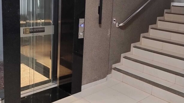 A modern hotel lobby with a glass elevator, a staircase with a metal handrail, and a textured wall. - Executive Hotel (Lucas do Rio Verde)