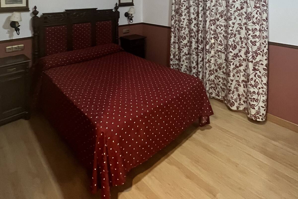 A bedroom with a wooden bed, a nightstand, a wall-mounted lamp, and a window with floral curtains.