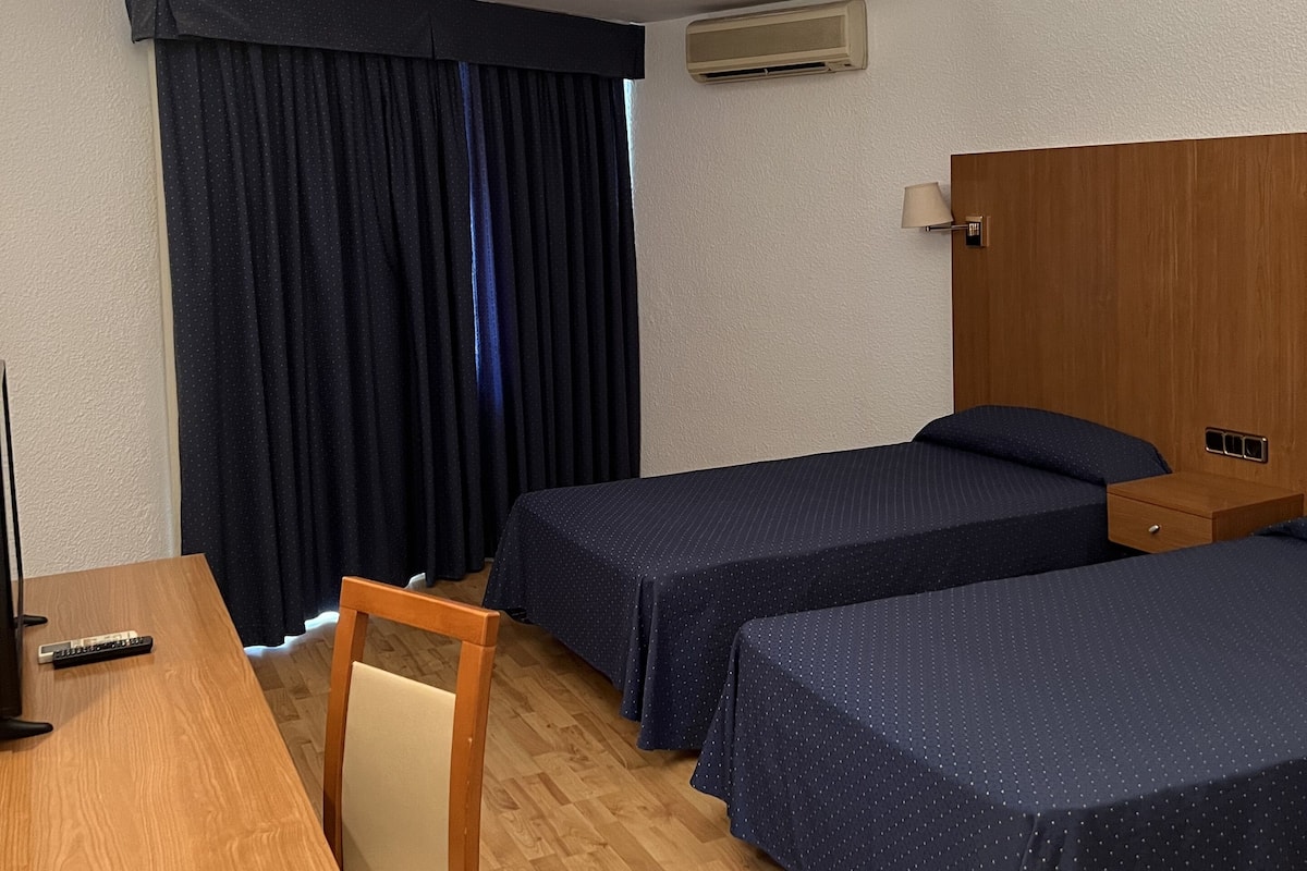 A hotel room with two single beds, a desk with a chair, a TV, and an air conditioning unit.
