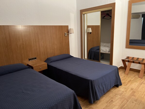 A hotel room with two single beds, a wooden headboard, a bedside table with a lamp, a mirror, and a wooden bench. - Hotel El Cisne (Zaragoza)