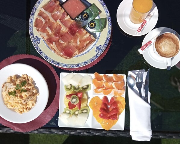 A table with a plate of prosciutto, a plate of fruit, a plate of scrambled eggs, a cup of coffee, and a juice.