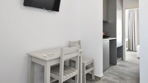 A modern, minimalistic room with a flat-screen TV mounted on the wall, a small white desk with a chair, and a kitchen area visible through an open doorway. - Imeres Studios & Apartments (Kassandra)