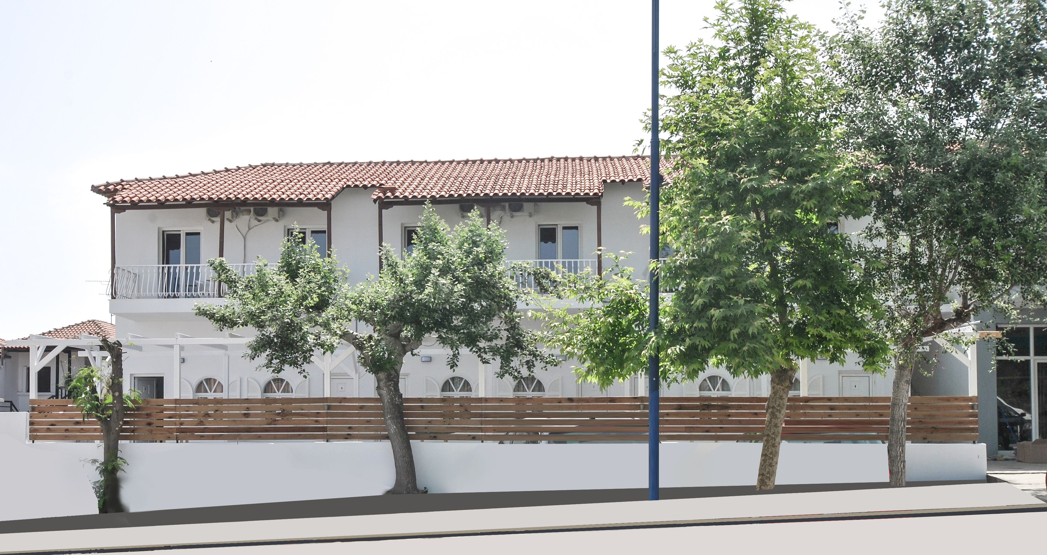 A two-story building with a tiled roof, white walls, and a balcony. There are trees in front of the building and a bench along the wall.
