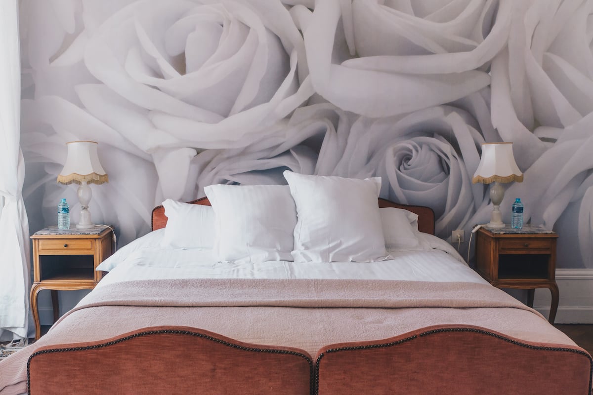 A bedroom with a bed, pillows, a nightstand, a lamp, and a wall with a floral mural.
