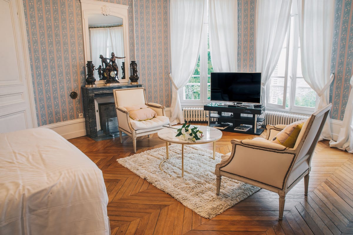 A room with a bed, two armchairs, a coffee table, a fireplace, a television, and a window with curtains.