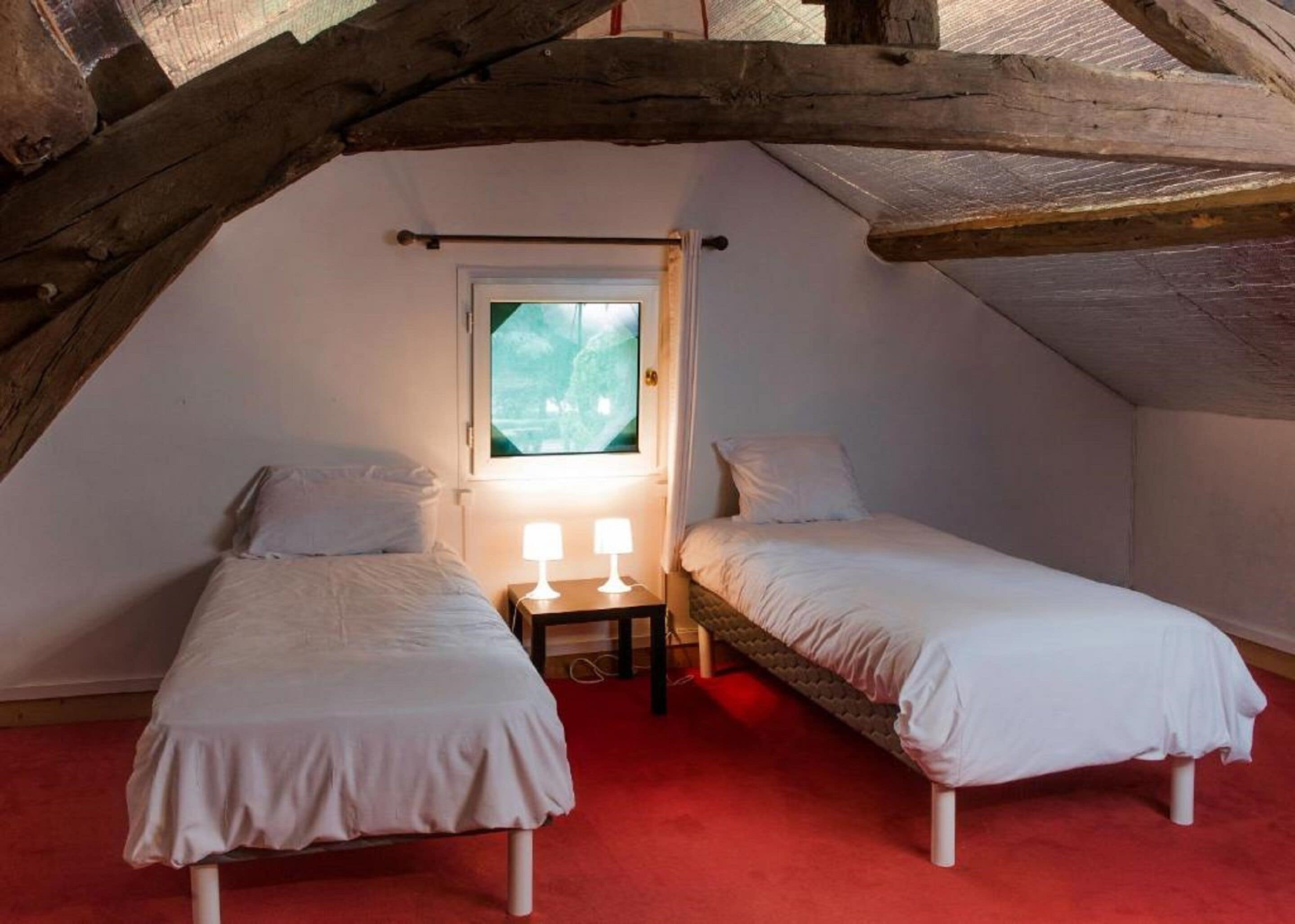 Une petite chambre mansardée, chaleureuse et accueillante, avec deux lits simples, une fenêtre et une moquette rouge.