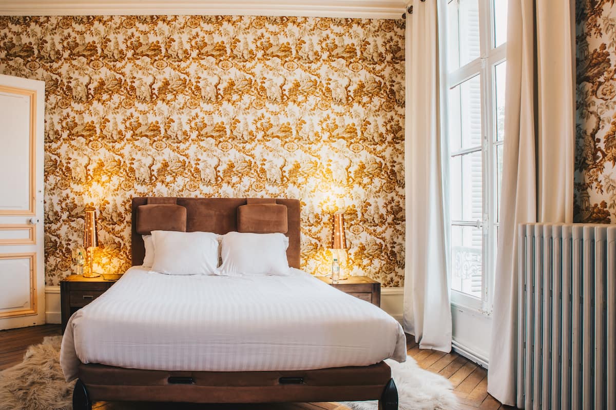 A bedroom with a large bed, wooden headboard, and patterned wallpaper. There is a window with curtains, a radiator, and a nightstand with a lamp.