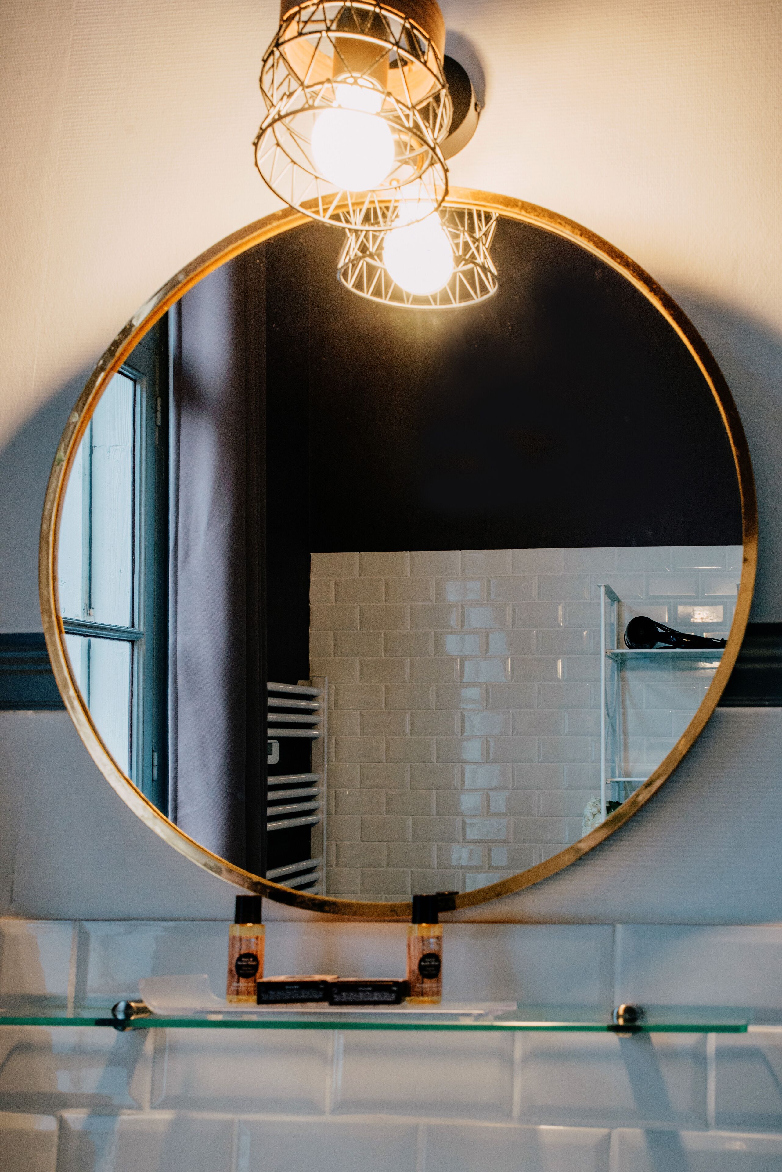 A round mirror with a gold frame mounted on a wall above a glass shelf.