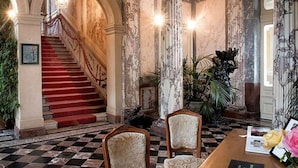 A grand staircase leading to an upper level, a checkered floor, and two upholstered chairs in a room with marble walls and columns.
