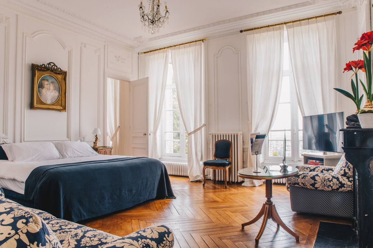 A hotel room with a bed, a sofa, a chair, a table, a TV, and a window with curtains.