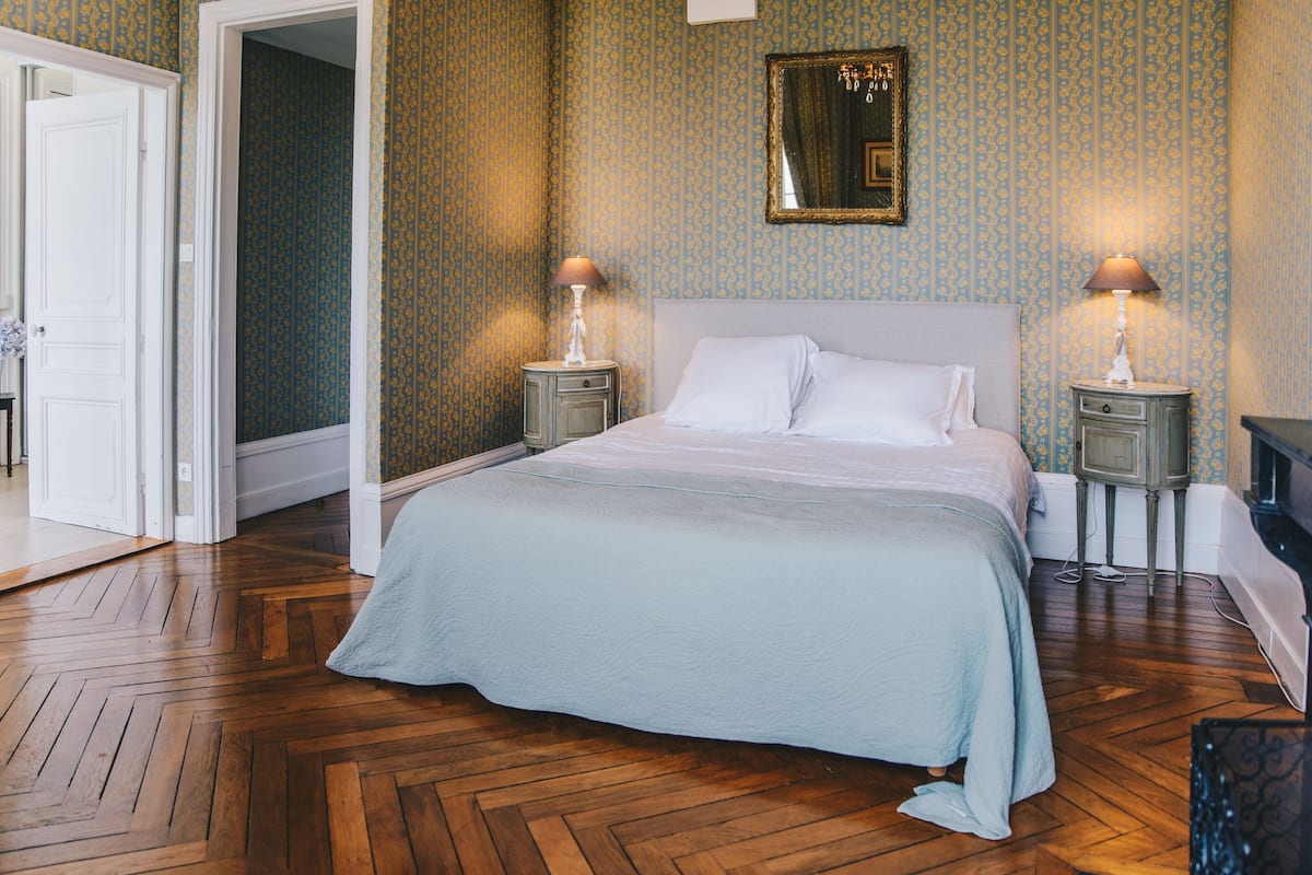 A bedroom with a large bed, two bedside tables with lamps, a mirror on the wall, and patterned wallpaper.