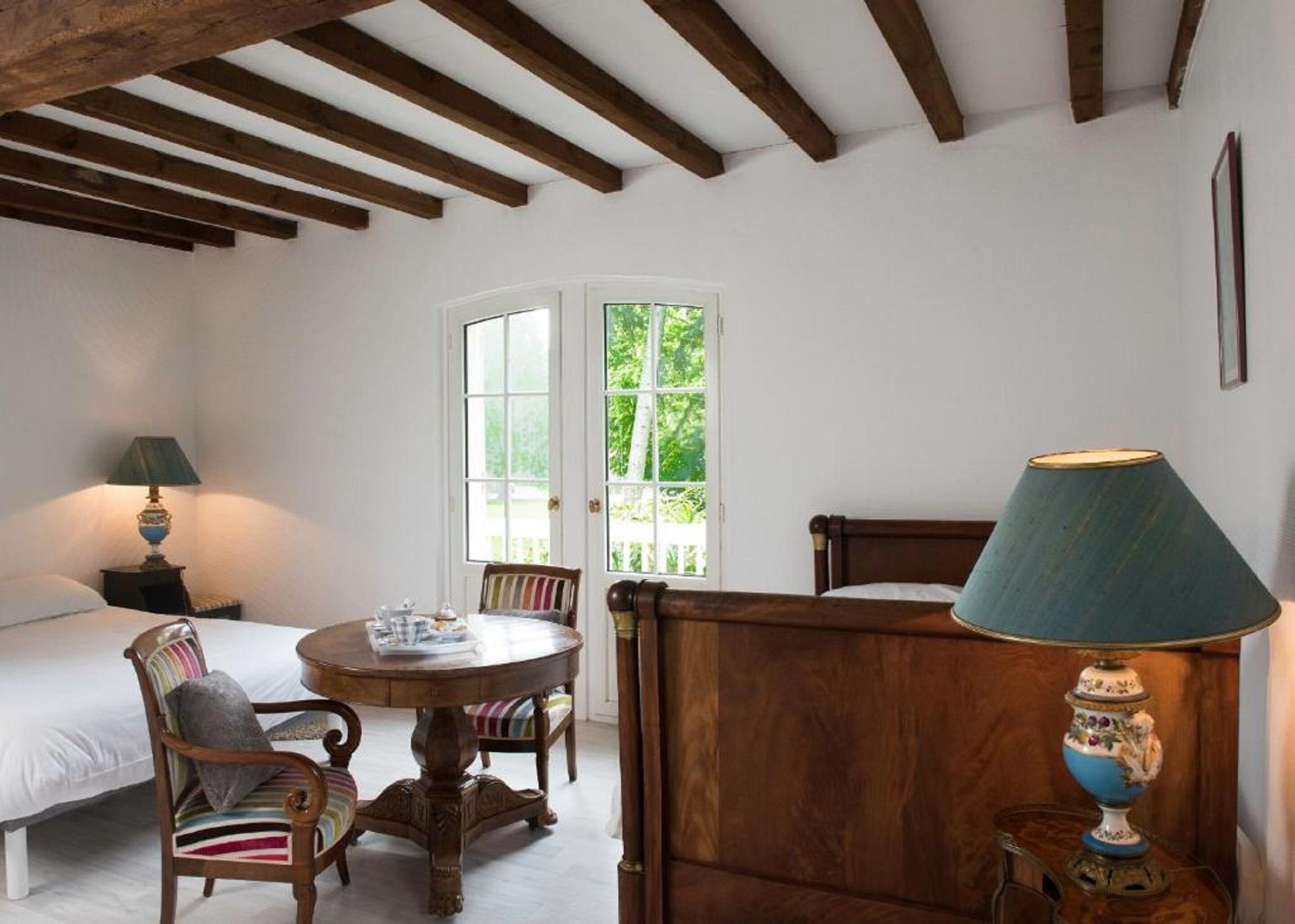 A room with a bed, a wooden chair, a small table, and a lamp. There is a window with a view of greenery outside.