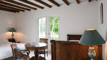 A room with a bed, a wooden chair, a small table, and a lamp. There is a window with a view of greenery outside.