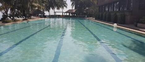 Una piscina con agua cristalina, rodeada de palmeras y un edificio con techo de paja.