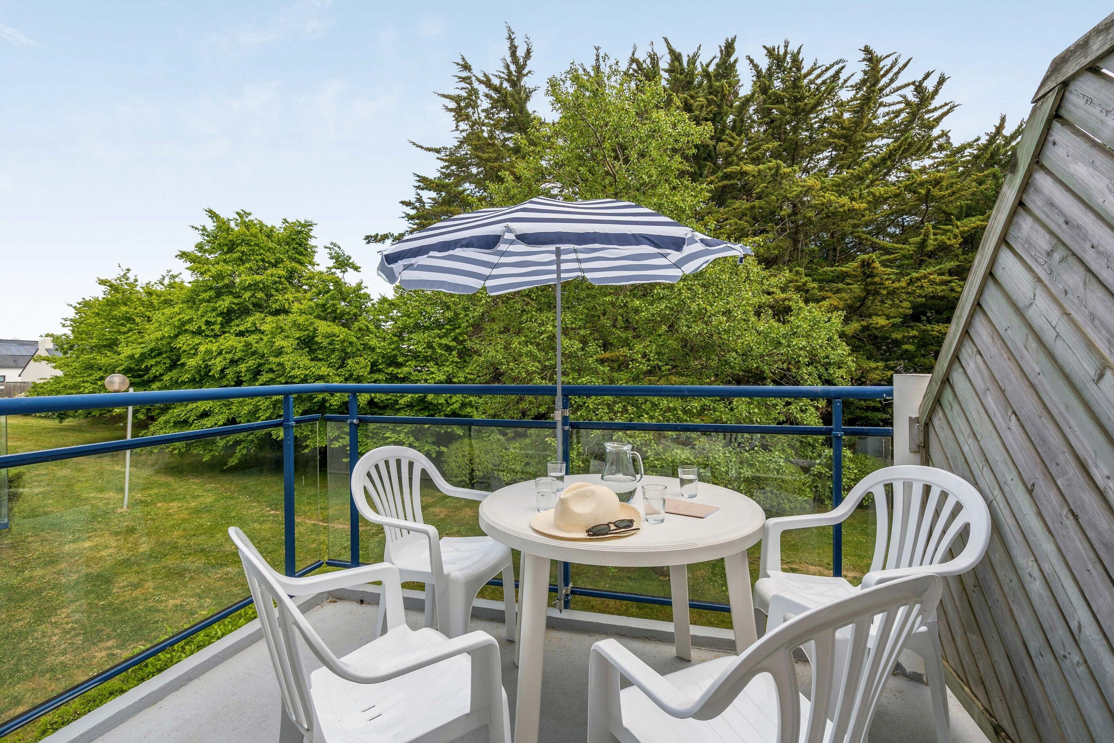 Un balcon avec une table et des chaises, un parasol à rayures et une vue sur la verdure environnante.