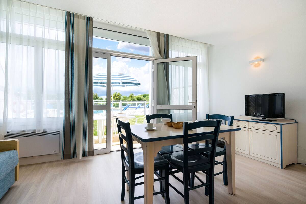 Une chambre moderne équipée d’une table à manger, de chaises, d’un téléviseur et offrant une vue sur un immeuble grâce à de grandes fenêtres.